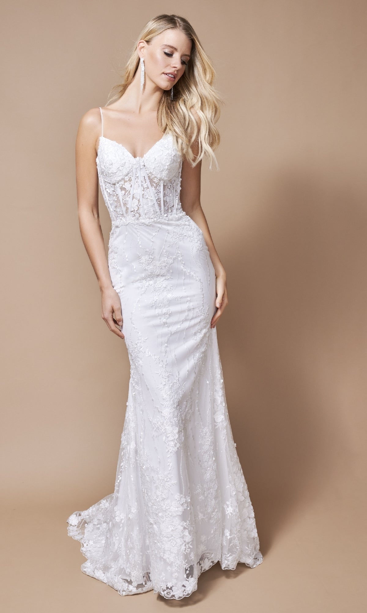 A woman in the Nina Canacci 2392 long formal dress, featuring a fitted lace design with thin straps and a sheer bodice, stands against a plain beige background.